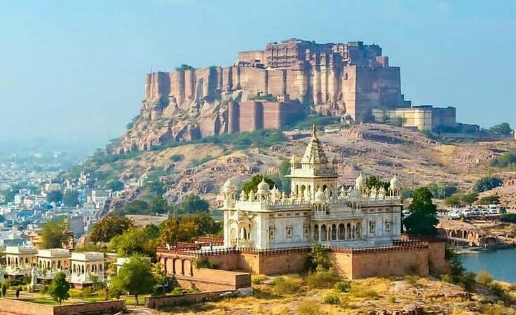 A stunning view of the majestic Jaisalmer Fort, perched on a sandy hill, under the clear blue sky. In the foreground, a beautiful white-domed temple stands elegantly, surrounded by the golden landscape of Jaisalmer. This picturesque scene captures the essence of the city, making it a perfect destination to explore with a reliable Jaisalmer Taxi Service.